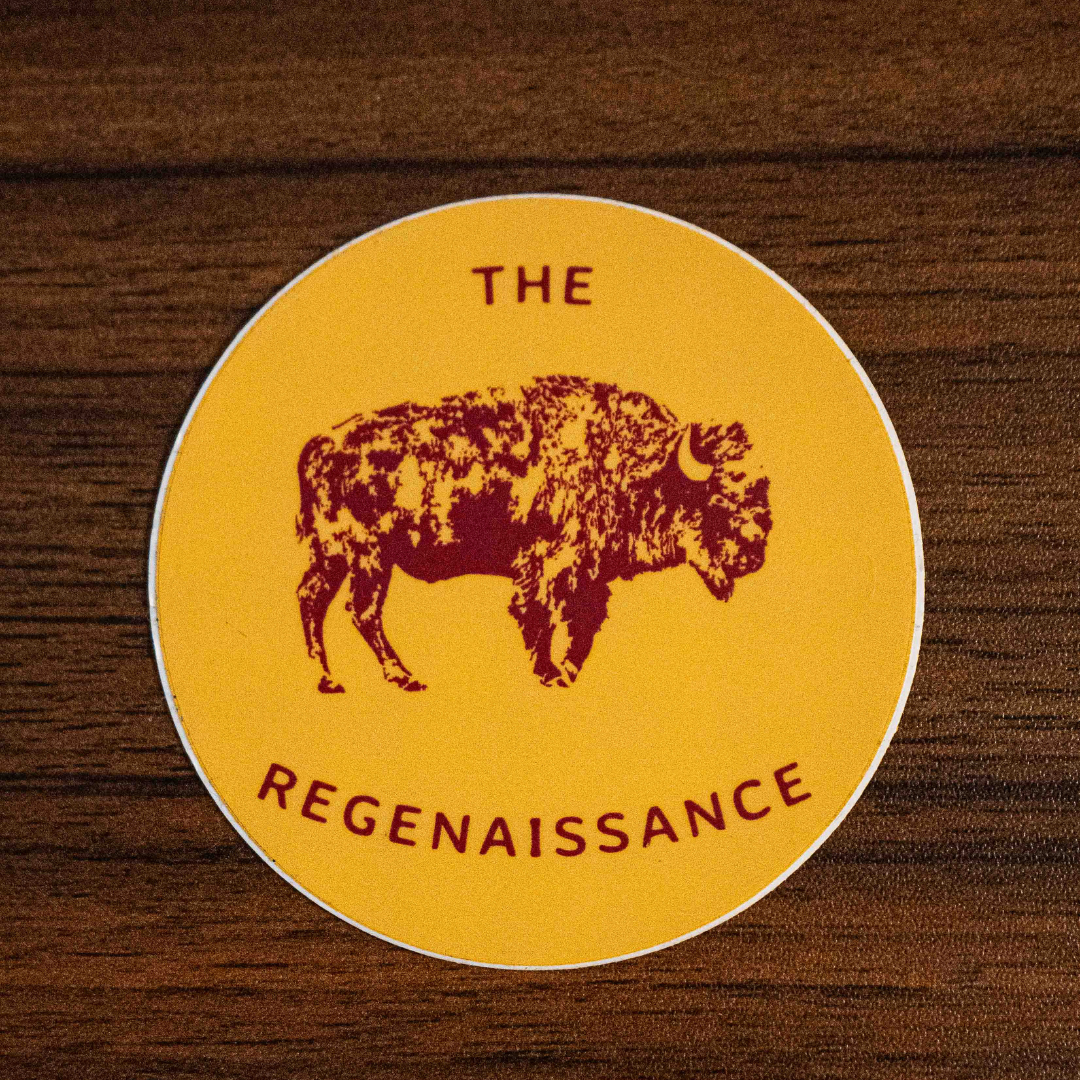 The Regenaissance Sticker by Sticker Mule is a round yellow vinyl sticker with a maroon Great American Bison and THE REGENAISSANCE in maroon, shown on a wooden surface.