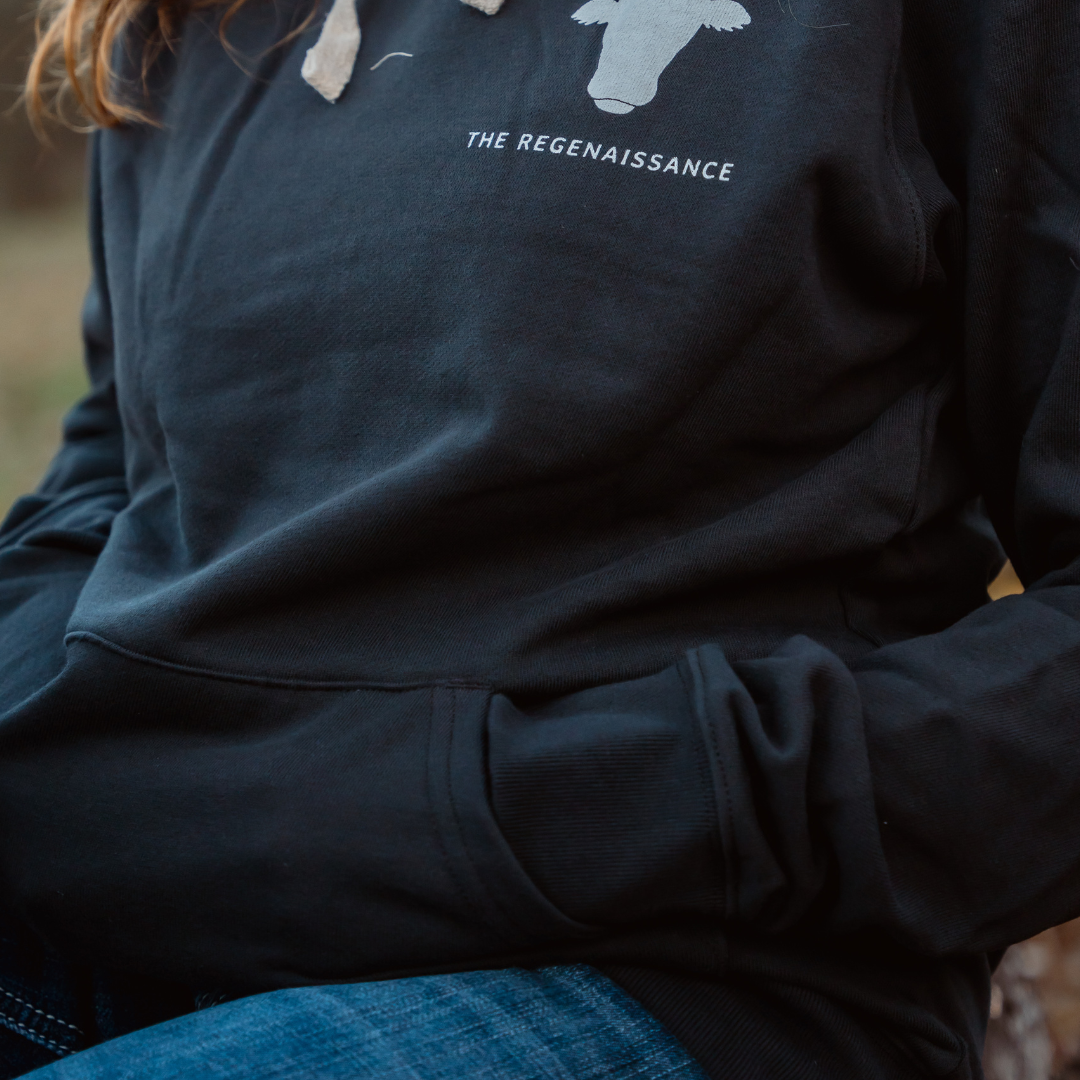 A person wears a Farm Fresh Clothing Raw is Law Hoodie with a bull head graphic and THE REGENAISSANCE text on the chest, paired with blue jeans. Hands in the pocket, they embody food freedom.