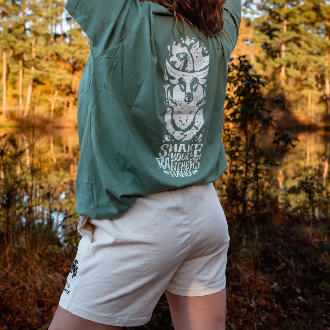A person stands outdoors with arms raised, wearing Los Angeles Apparel Womens Bison Shorts and a green graphic T-shirt. Trees and a lake are in the background, evoking a natural setting inspired by regenerative agriculture.