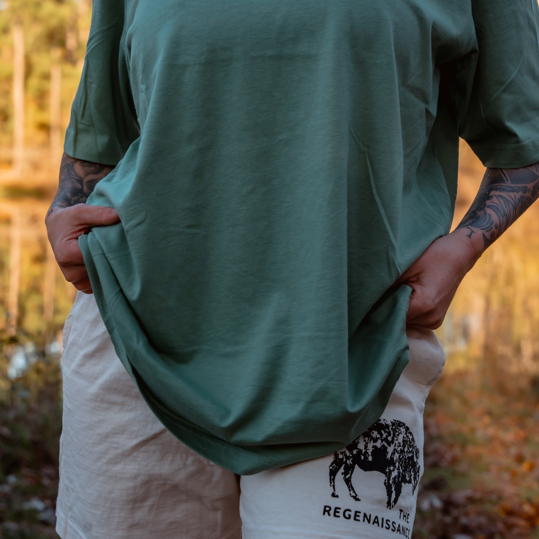 A person stands outdoors wearing a green THE REGENAISSANCE t-shirt and Los Angeles Apparel Womens Bison Shorts in creme, inspired by regenerative agriculture. Trees and sunlight enhance the background.