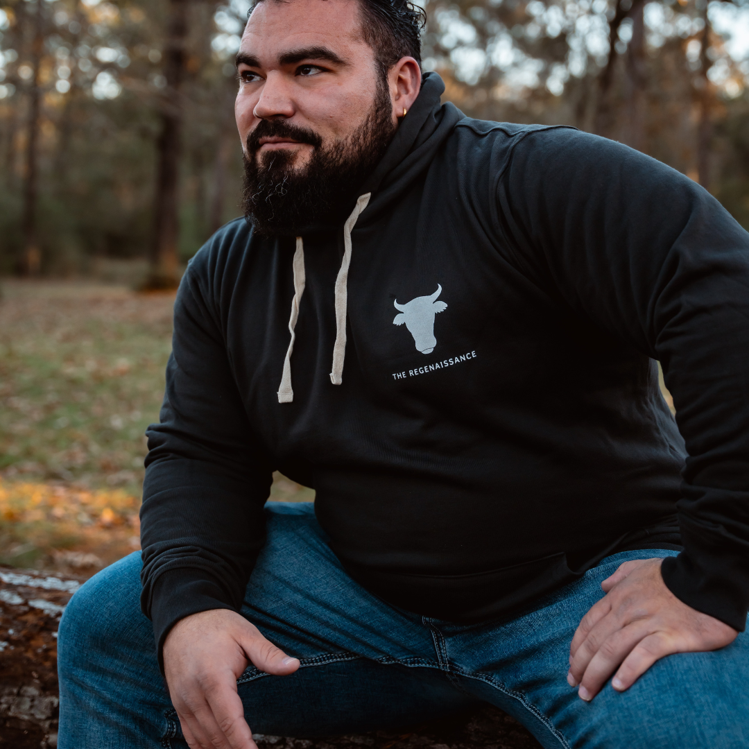 A bearded man in a gunpowder Raw is Law Hoodie from Farm Fresh Clothing and blue jeans sits on a fallen log outdoors, relaxed and enjoying nature’s peace and the spirit of food freedom as sunlight filters through the forest behind him.