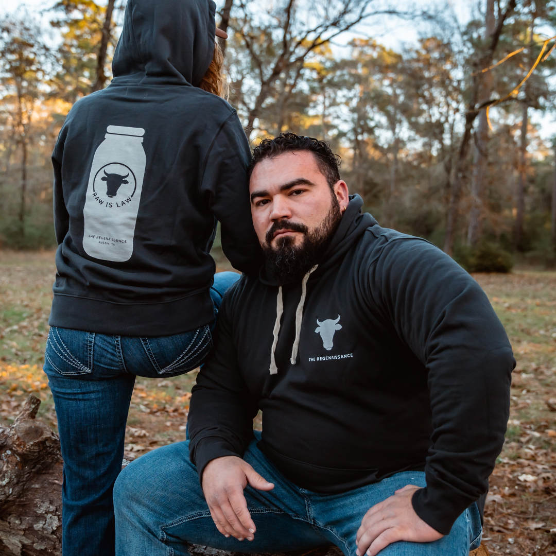 A man outdoors wears a black Farm Fresh Clothing hoodie with a white cow logo, ideal for food freedom and raw milk fans. Next to him, another person sports the Raw is Law Hoodie featuring a jar graphic and the phrase “How Is Lunch?”.