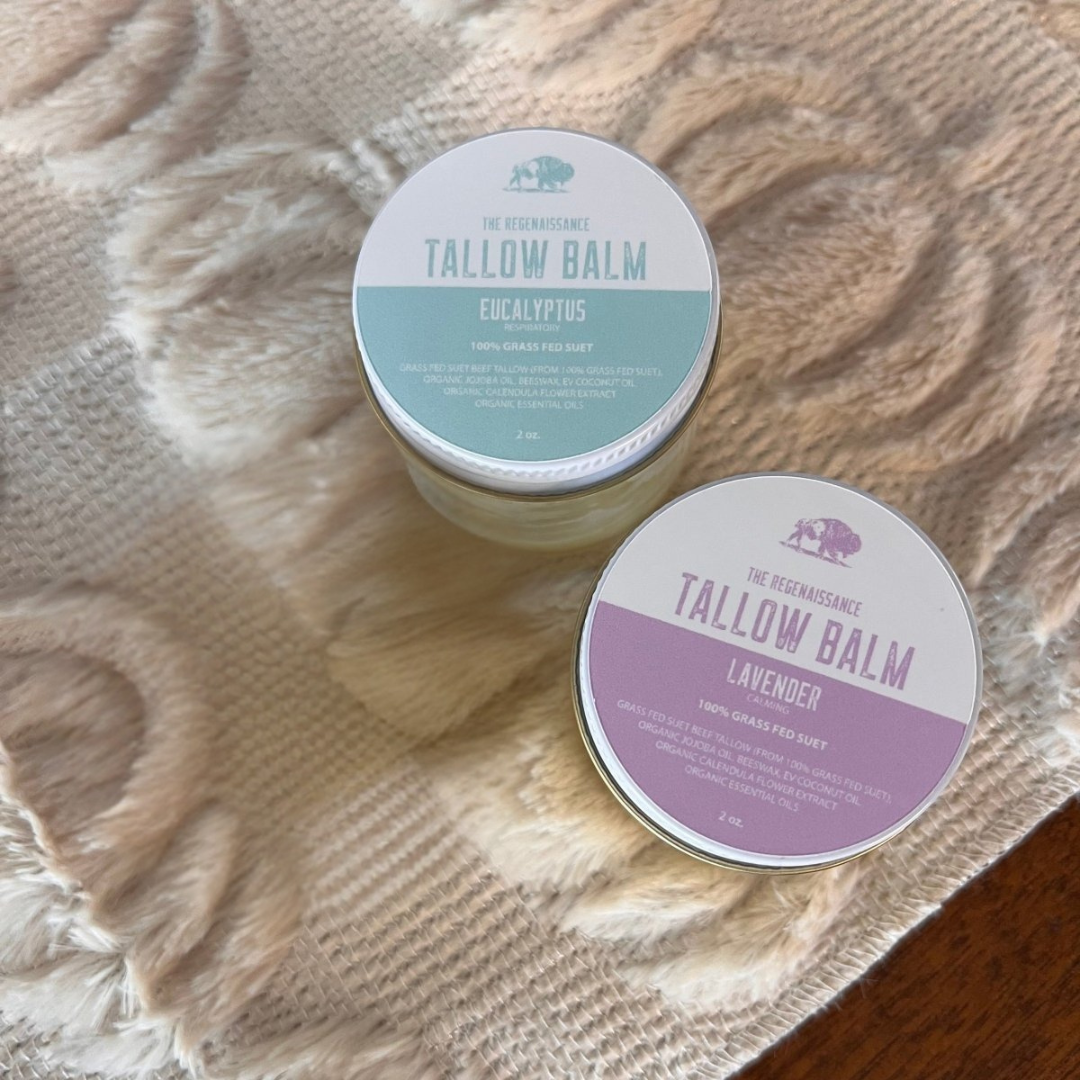 Two small round metal tins of Tallow Balm by The Regenaissance, one labeled Eucalyptus with a light blue label and the other labeled Lavender with a purple label, placed on a beige textured blanket over a brown wooden surface.