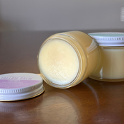 Two small jars of balm on a wooden surface, with one jar tipped over to show the pale yellow balm inside and the lid placed beside it.