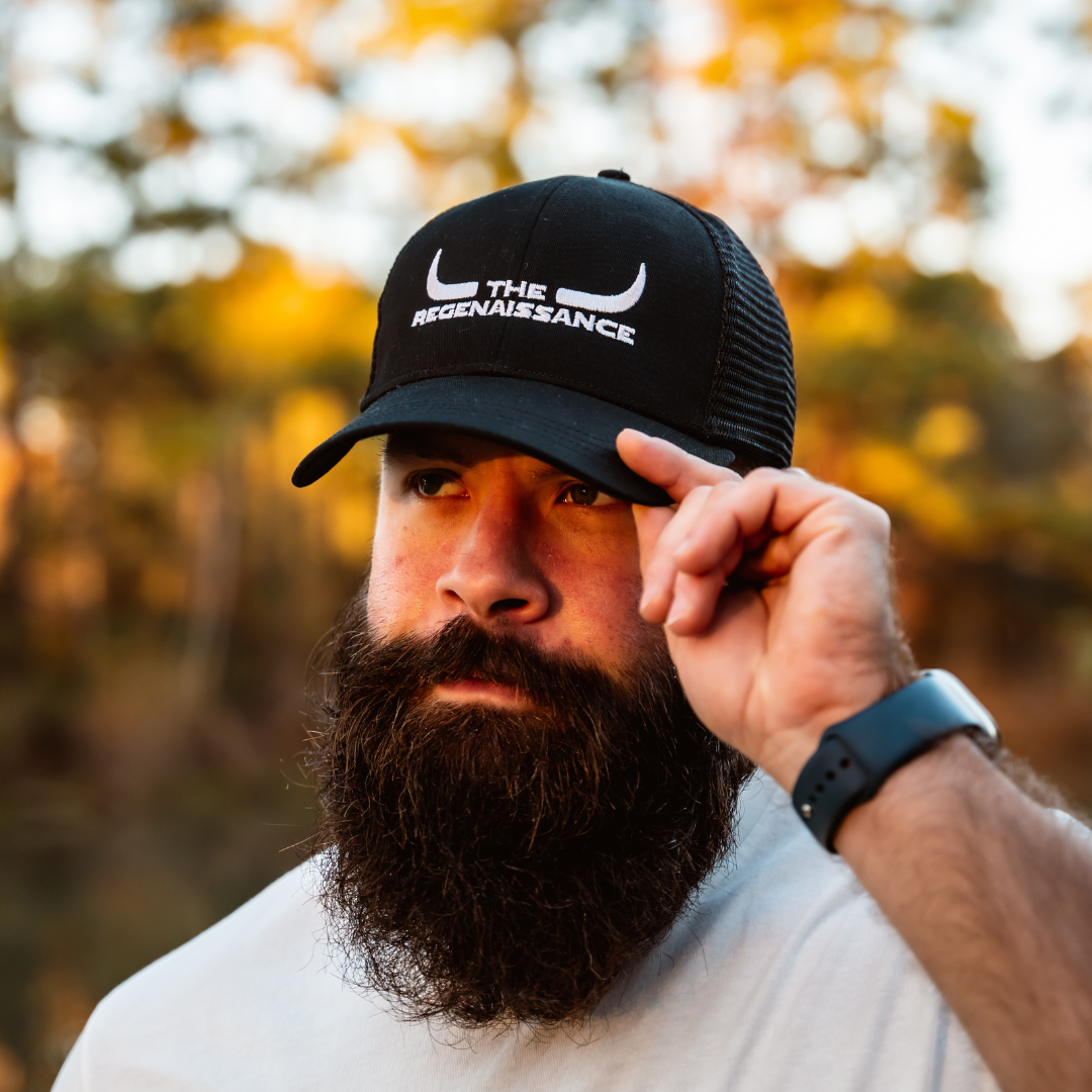 black Longhorn Trucker Hat from Farm Fresh Clothing stands outdoors, holding the brim of his hat. Trees and sunlight in the blurry background evoke the spirit of food freedom.