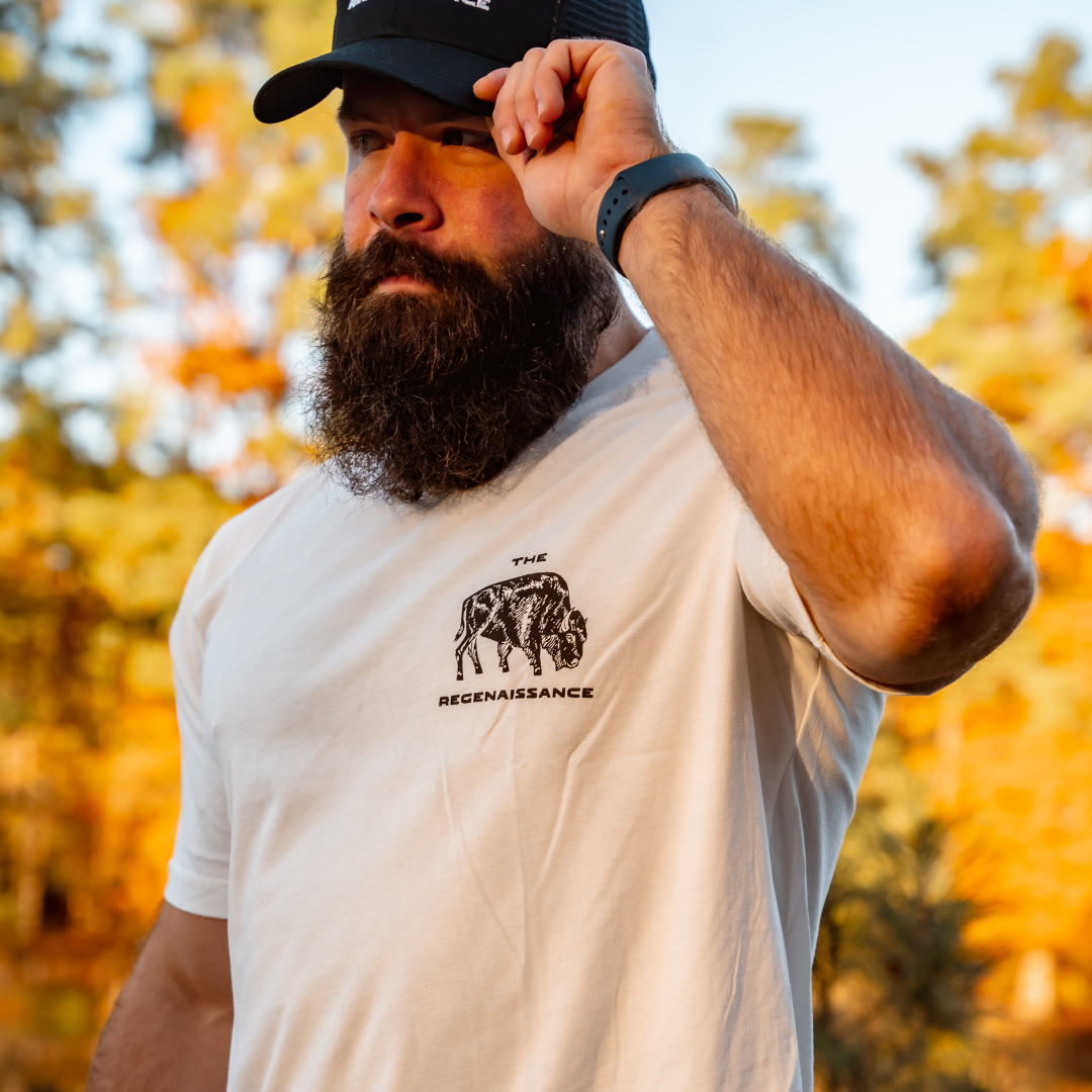 A bearded man in a white tee featuring a bull and The Regenaissance adjusts his Farm Fresh Clothing Longhorn Trucker Hat outdoors, with blurred autumn trees and yellow leaves in the background.