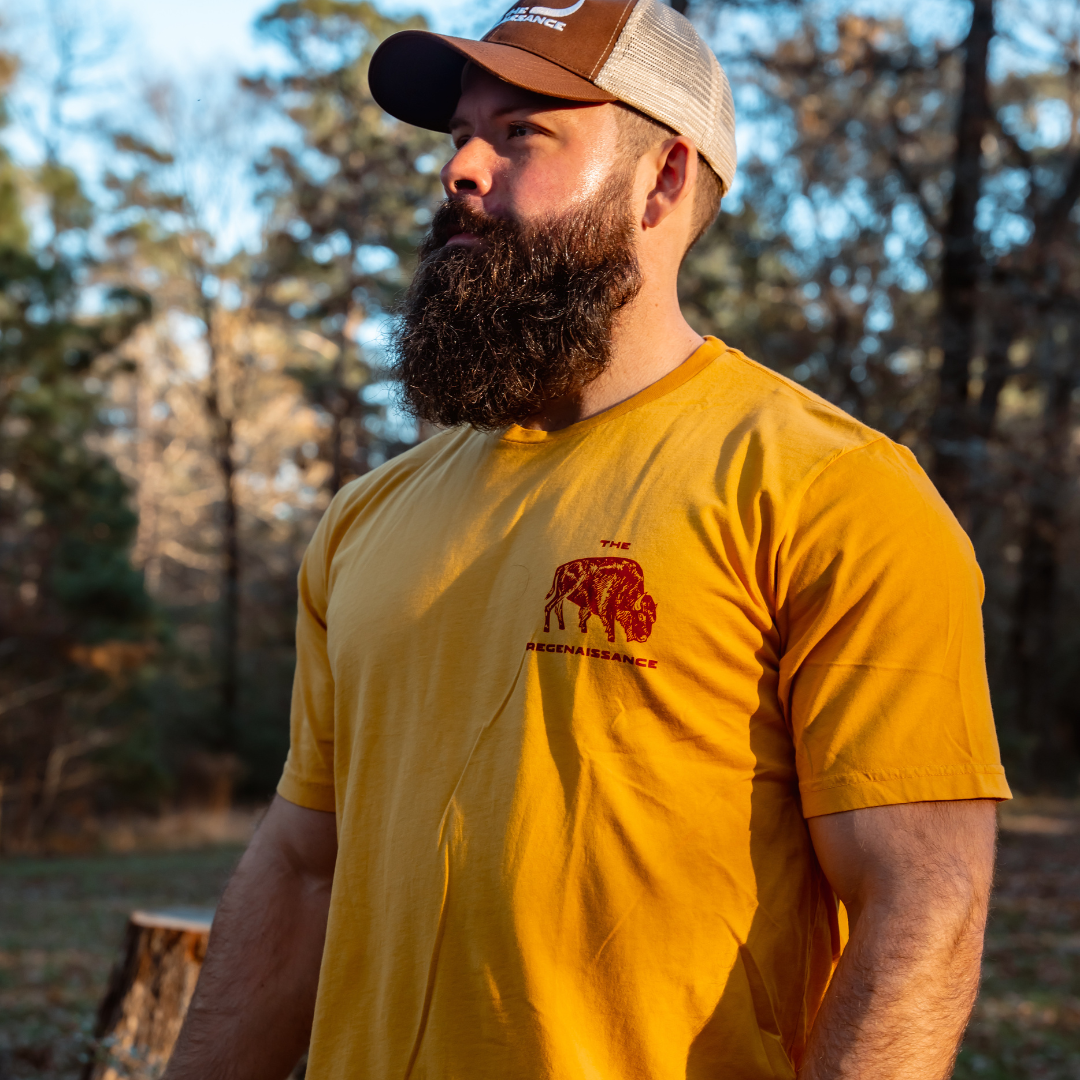 A bearded man in a yellow T-shirt and Farm Fresh Clothing’s Longhorn Trucker Hat stands outdoors among sunlit autumn trees, evoking a sense of food freedom and connection to nature.