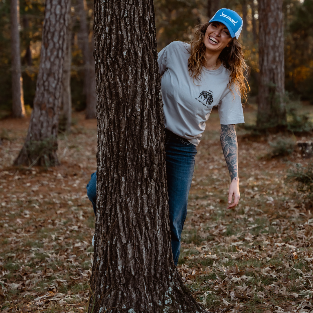 A smiling woman in a blue Farm Fresh Clothing Longhorn Trucker Hat, gray tee, and jeans playfully leans out from behind a tree amid fallen leaves, embracing food freedom.