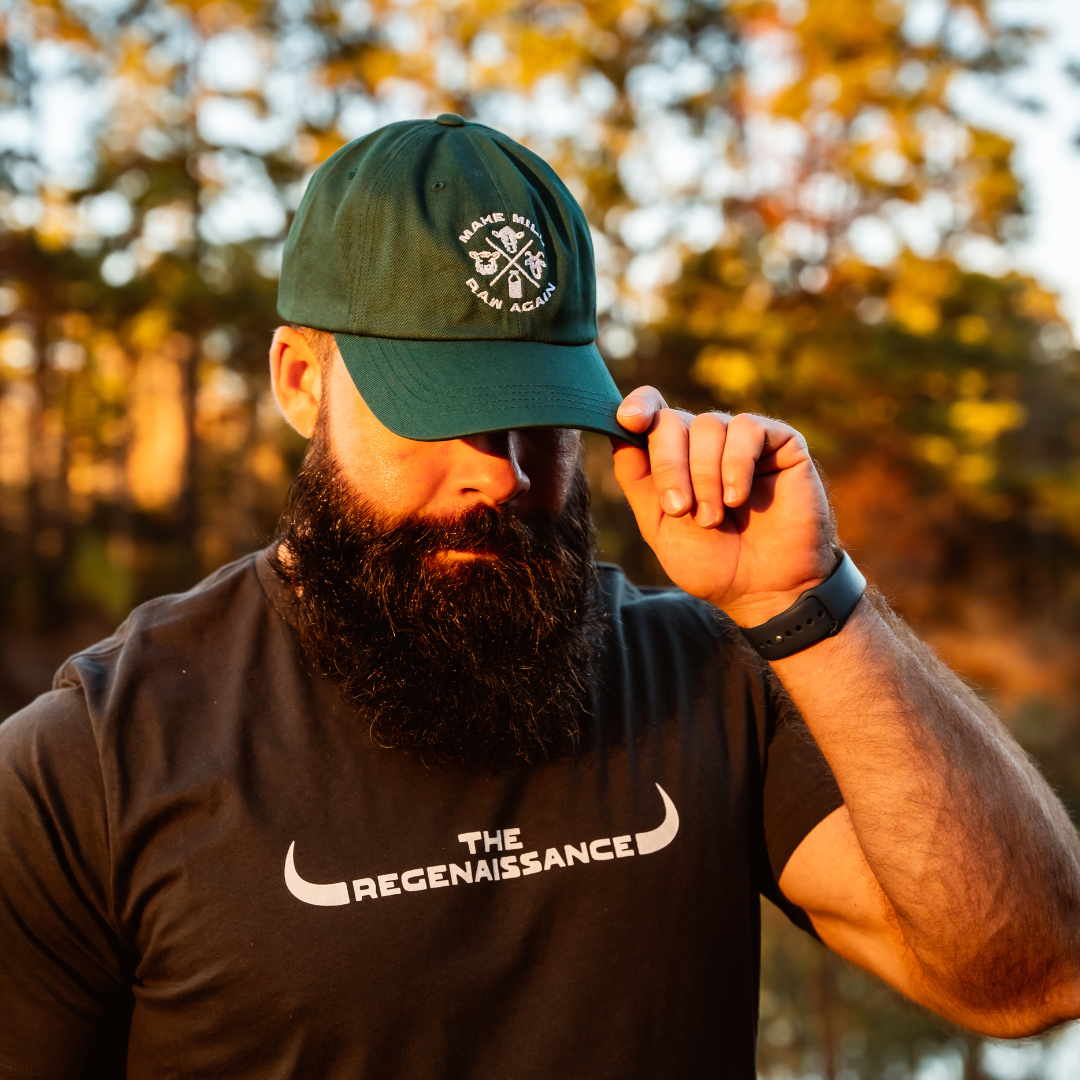 A bearded man in a spruce MMRA Trinity Cap by Farm Fresh Clothing and a The Regenaissance t-shirt stands outdoors, tilting his cap downward amid trees and warm sunlight, capturing the essence of food freedom.