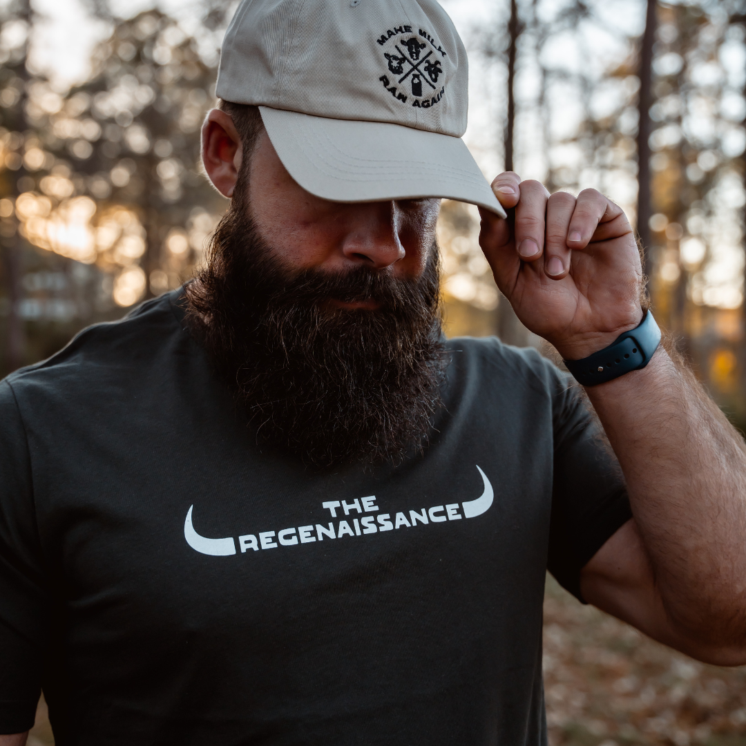 A bearded man outdoors wears a dark shirt and a light stone MMRA Trinity Cap by Farm Fresh Clothing, holding the cap’s brim with one hand. Blurred trees are visible in the background.