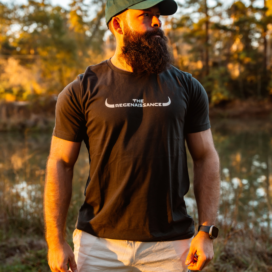 A bearded man in a dark THE REGENAISSANCE tee, white shorts, and a Farm Fresh Clothing MMRA Trinity Cap stands outdoors by a pond amid trees and soft golden sunlight.