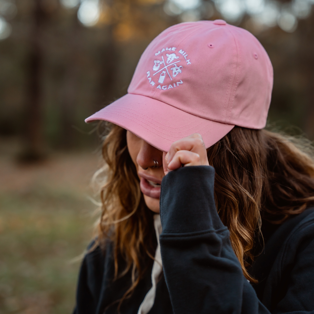 A wavy-haired person with a nose ring wears a pink Farm Fresh Clothing MMRA Trinity Cap and a dark hoodie, tilting the cap to partially hide their face while standing outdoors in a blurred wooded area.