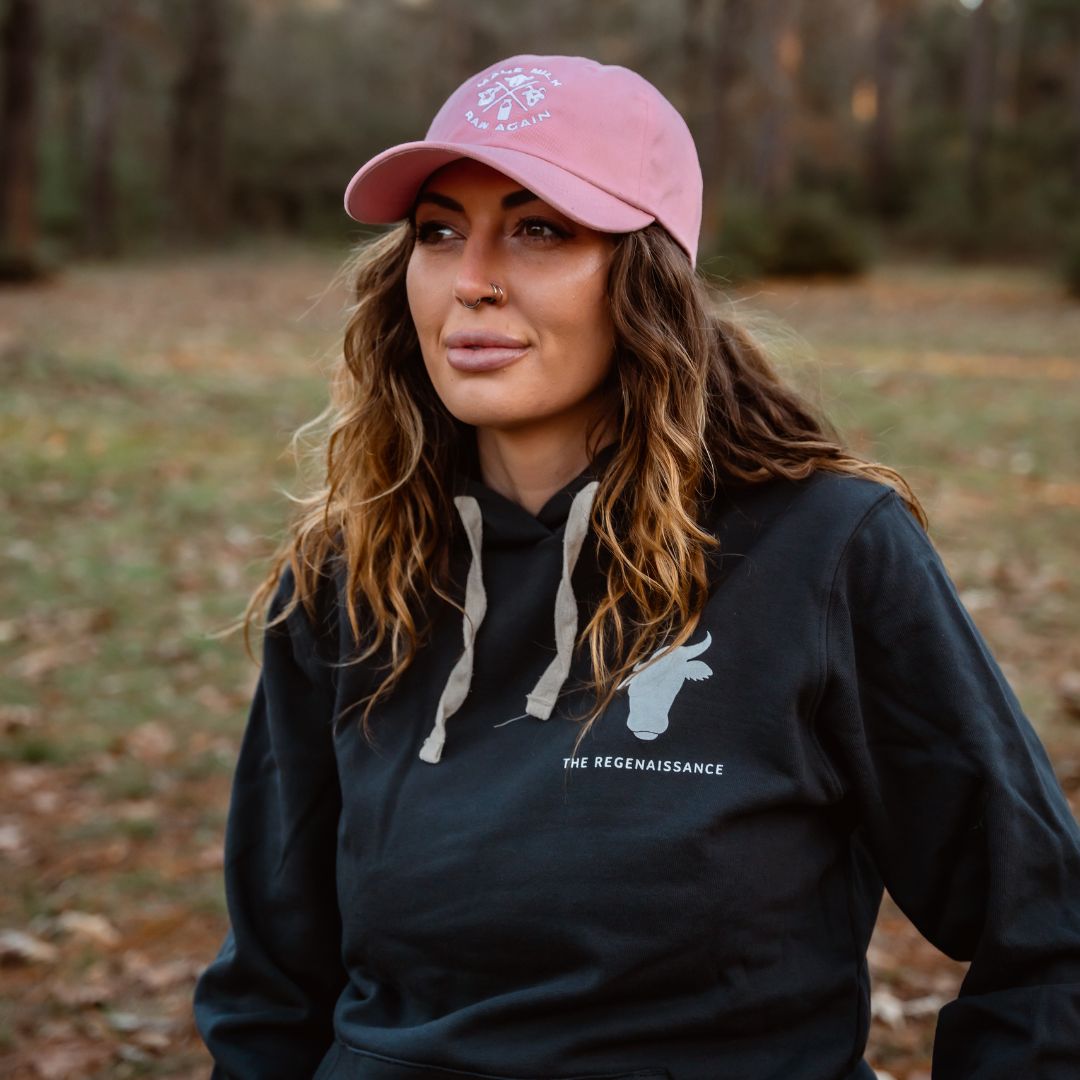 A woman with wavy brown hair sits outdoors in a Farm Fresh Clothing MMRA Trinity Cap and a black hoodie with THE REGENAISSANCE text. Blurred trees and grass in the background evoke food freedom.