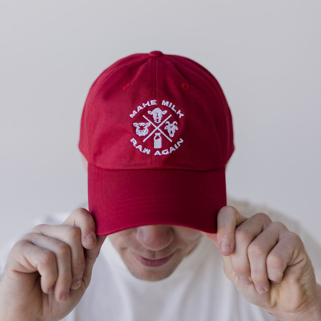 Wearing a white shirt, someone holds the brim of the cranberry MMRA Trinity Cap by Farm Fresh Clothing, embroidered with “MAKE MILK RAW AGAIN” and white icons—showing support for food freedom.