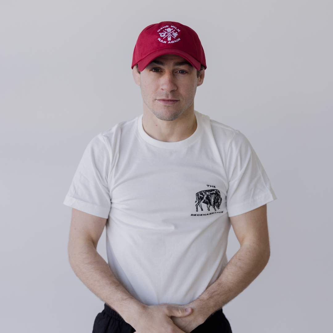 Against a plain light background, a person wears a red Farm Fresh Clothing MMRA Trinity Cap, a white tee with small black chest graphic, and black pants. Their hands are loosely clasped in front, embodying the spirit of food freedom.