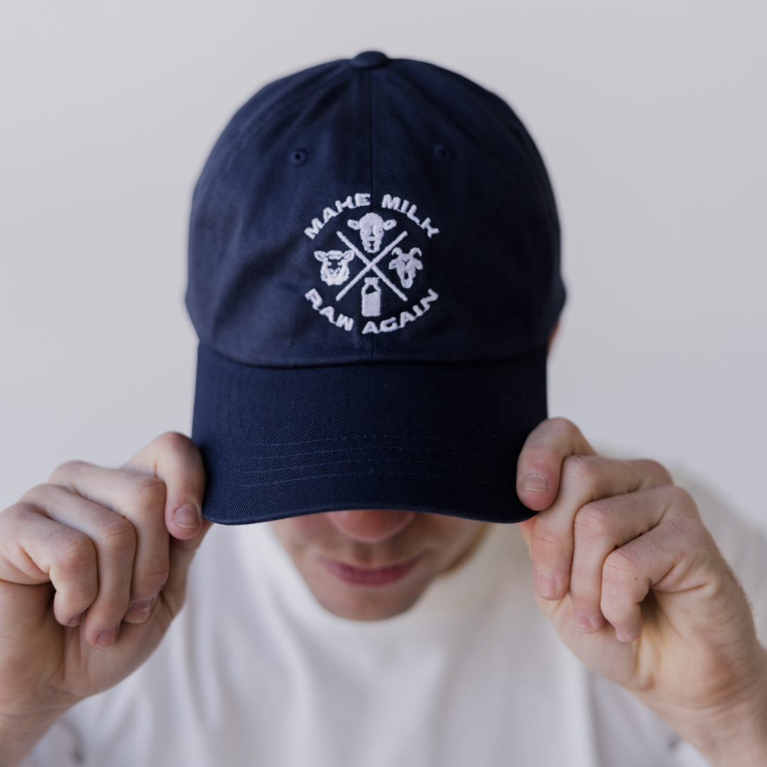 A person in a white shirt adjusts a navy MMRA Trinity Cap by Farm Fresh Clothing, featuring MAKE MILK RAW AGAIN and dairy images. Their face is mostly obscured by the brim, highlighting support for raw milk and food freedom.