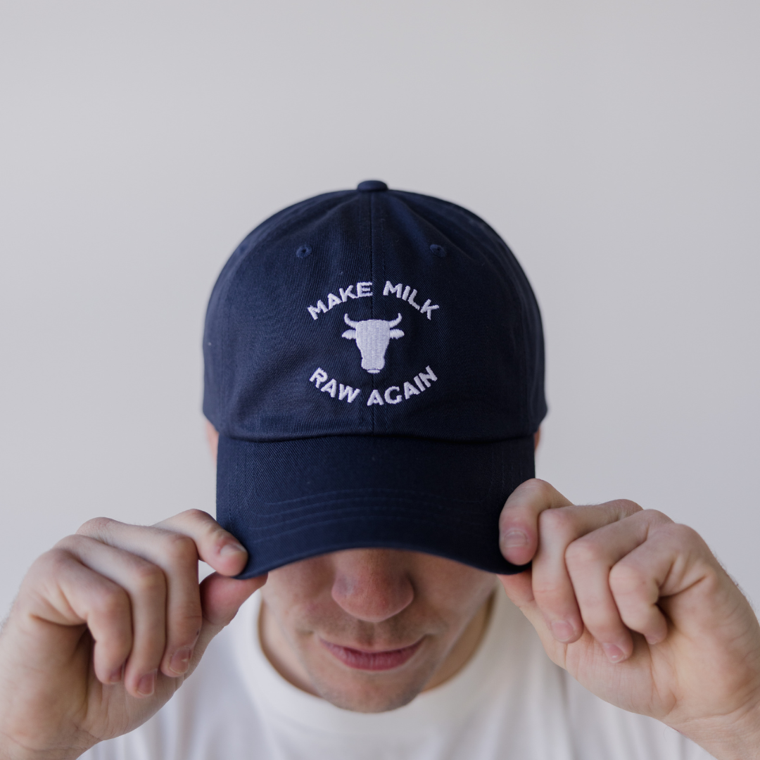 Wearing the navy MMRA Cow Cap by Farm Fresh Clothing—embroidered with a cow head and “MAKE MILK RAW AGAIN”—someone holds the brim, nodding to food freedom and raw milk advocacy against a light background.