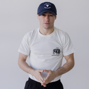 A man stands against a plain light background, wearing the Farm Fresh Clothing MMRA Cow Cap, a white t-shirt with a small chest graphic, and black pants, with his hands held together in front of him.