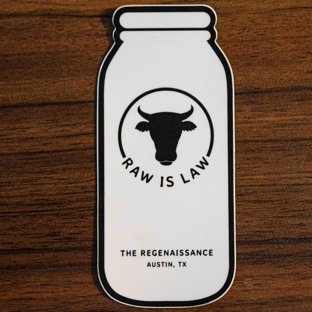 The Raw Is Law Sticker by Sticker Mule features a milk bottle with a cows head and RAW IS LAW text, plus THE REGENAISSANCE AUSTIN, TX below. Shown resting on a wooden surface, it highlights food rights.