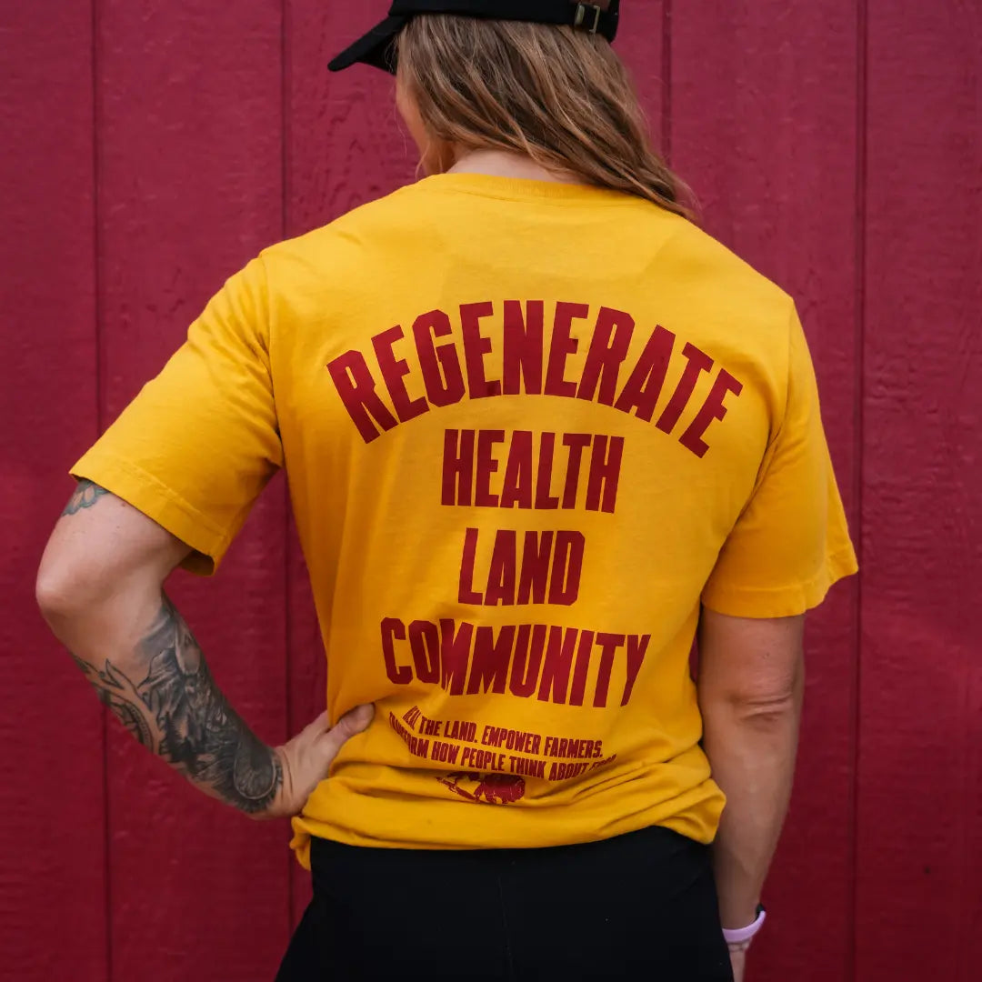 Woman wearing mustard Regenerate Agriculture Tee, back view, showing red “Regenerate Health Land Community” graphic against a red wall.