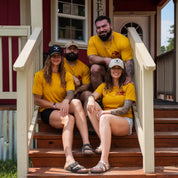 Group of two men and two women wearing mustard Regenerate Agriculture Tees with red bison logo, sitting together on farmhouse steps.