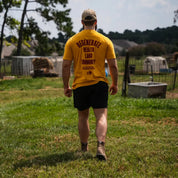 Man wearing mustard Regenerate Agriculture Tee, back view, showing bold red print, walking in a grassy farm field.