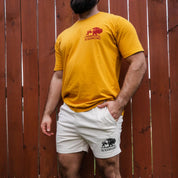 Man wearing mustard Regenerate Agriculture Tee, front view, paired with white Regenaissance Creme shorts, standing against wooden fence.