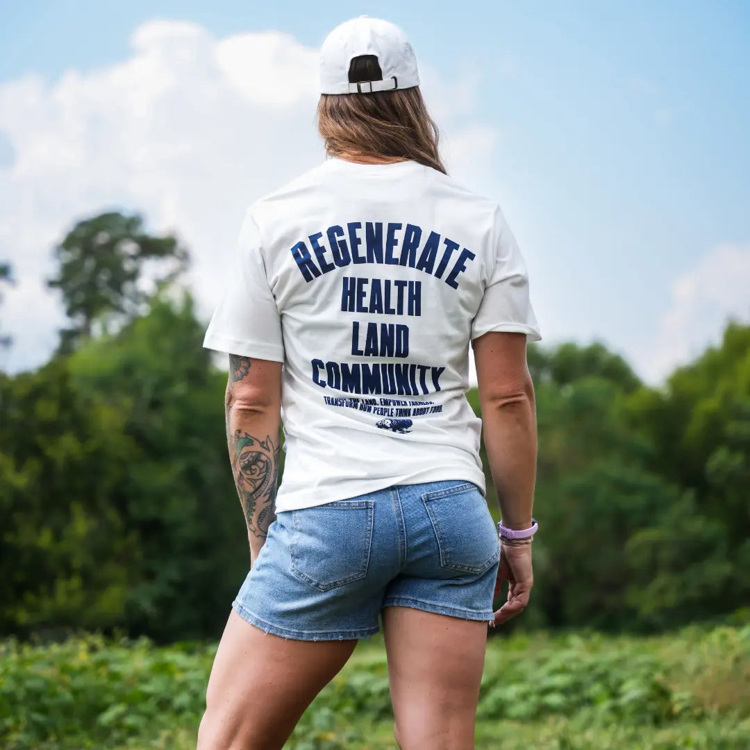 Woman wearing eggshell Regenerate Agriculture Tee, back view, showing navy “Regenerate Health Land Community” graphic while standing in a field.