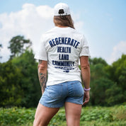 Woman wearing eggshell Regenerate Agriculture Tee, back view, showing navy “Regenerate Health Land Community” graphic while standing in a field.