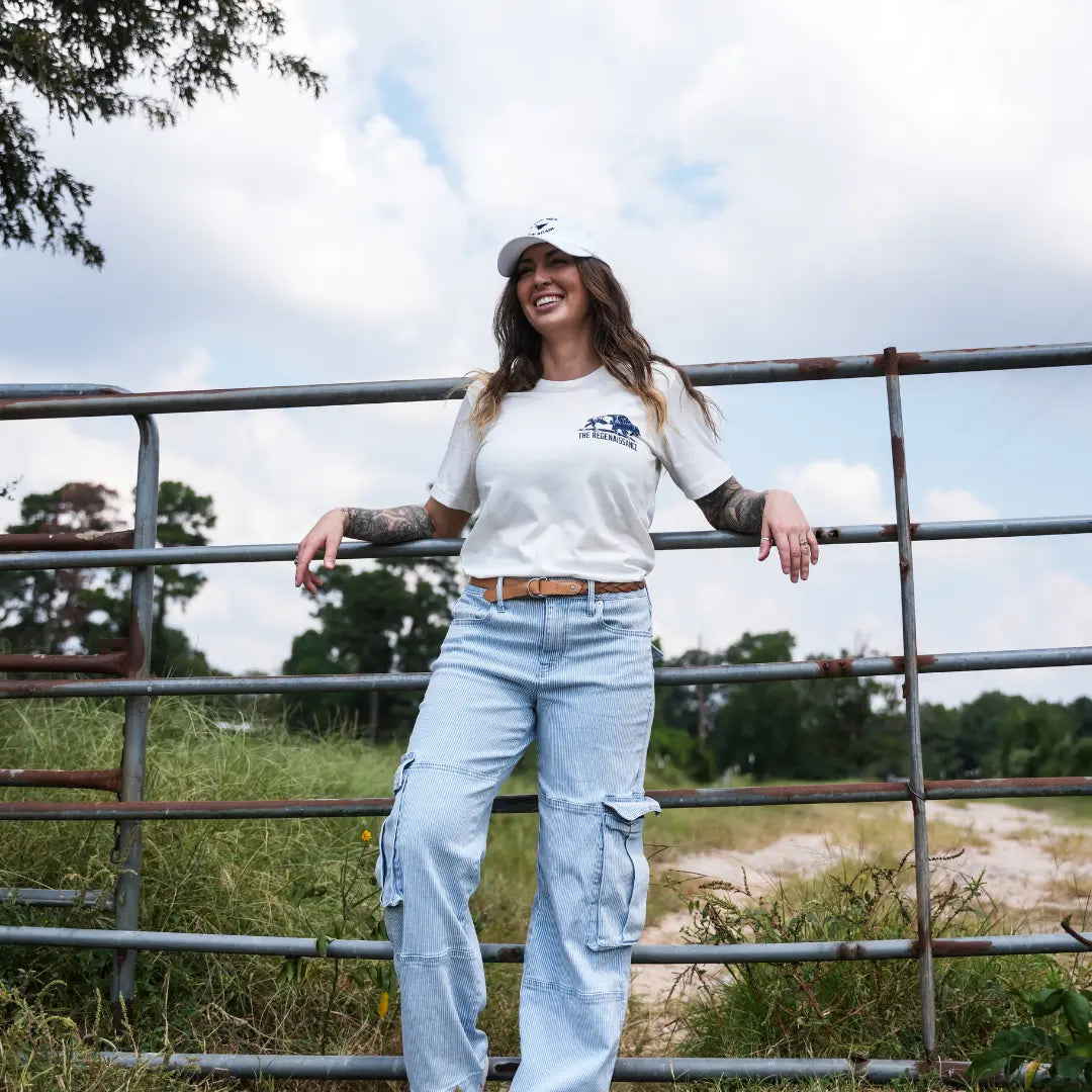 Woman smiling in eggshell Regenerate Agriculture Tee, front view, with navy bison and The Regenaissance logo, leaning on farm gate.