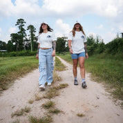 Two women walking on a dirt farm path wearing eggshell Regenerate Agriculture Tees with navy bison logo and The Regenaissance branding.