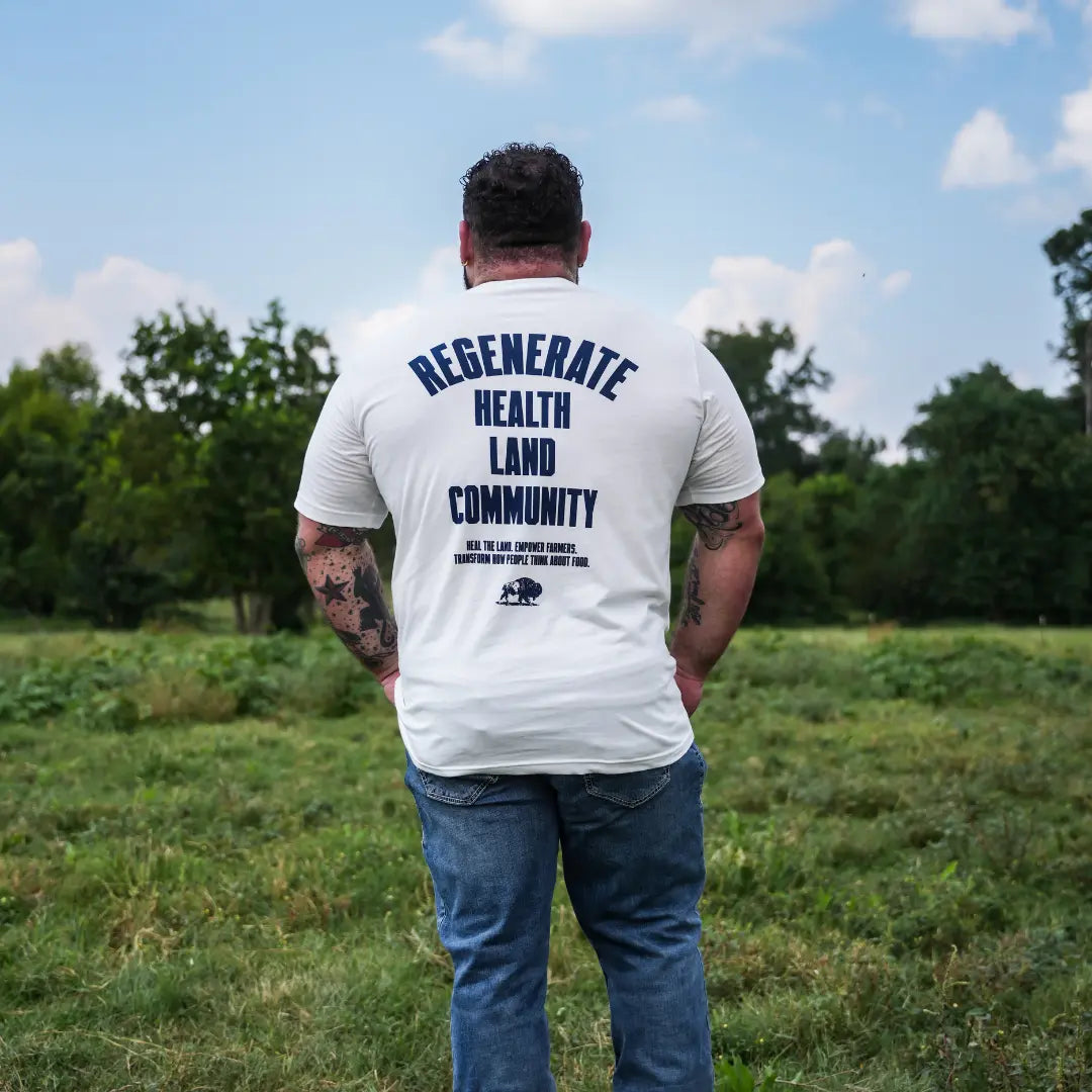 Man standing in grassy field wearing eggshell Regenerate Agriculture Tee, back view with bold navy “Regenerate Health Land Community” graphic.