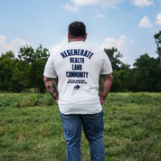 Man standing in grassy field wearing eggshell Regenerate Agriculture Tee, back view with bold navy “Regenerate Health Land Community” graphic.