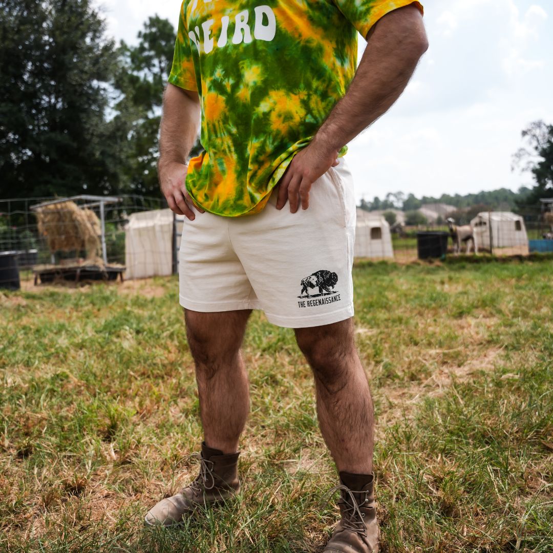 Side profile of male model wearing crème Regen Men’s Shorts, showing pocket depth and hem length on a raw milk farm.