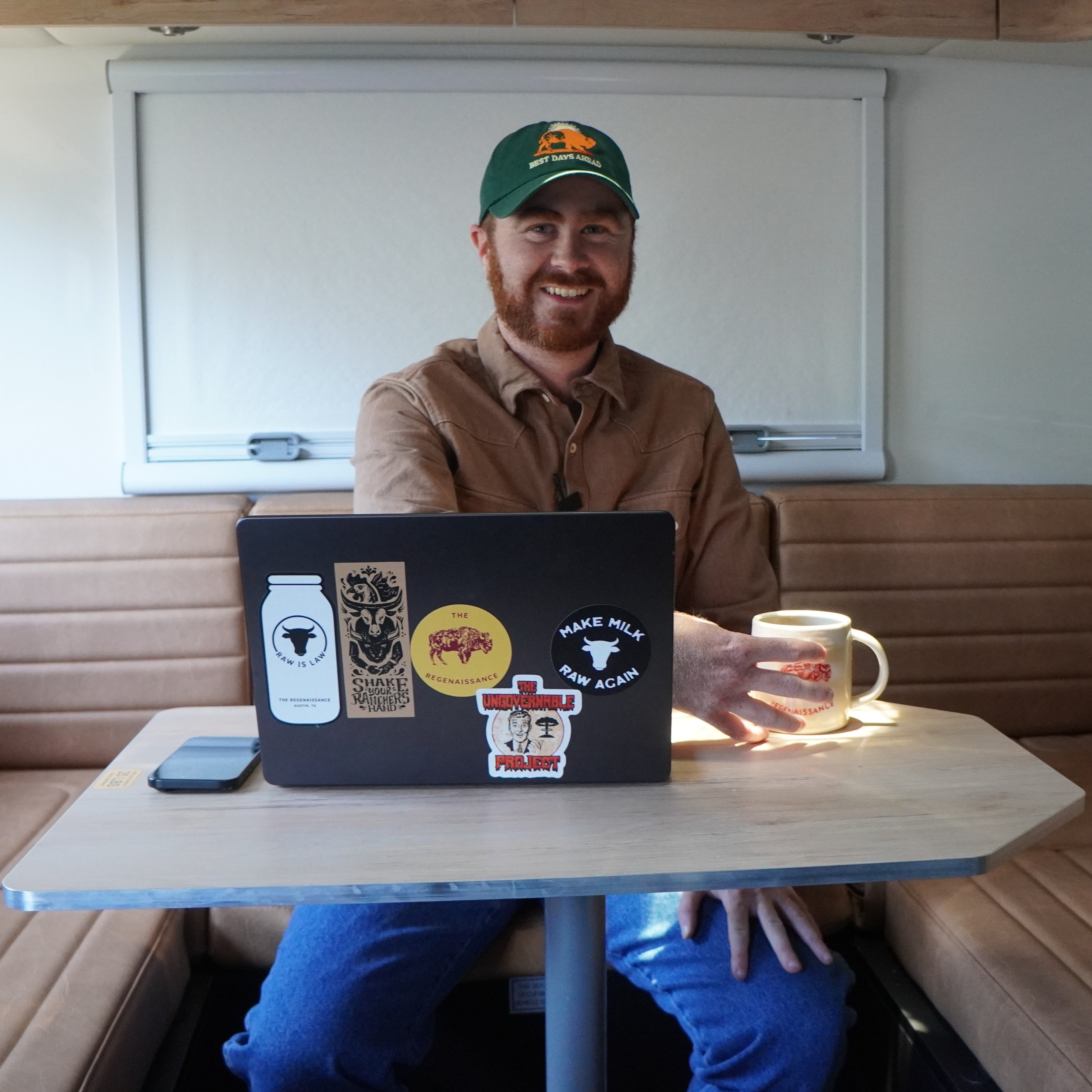 Ryan sitting inside a camper booth with a laptop covered in agricultural and food freedom stickers, smiling while holding a coffee mug with The Regenaissance logo.