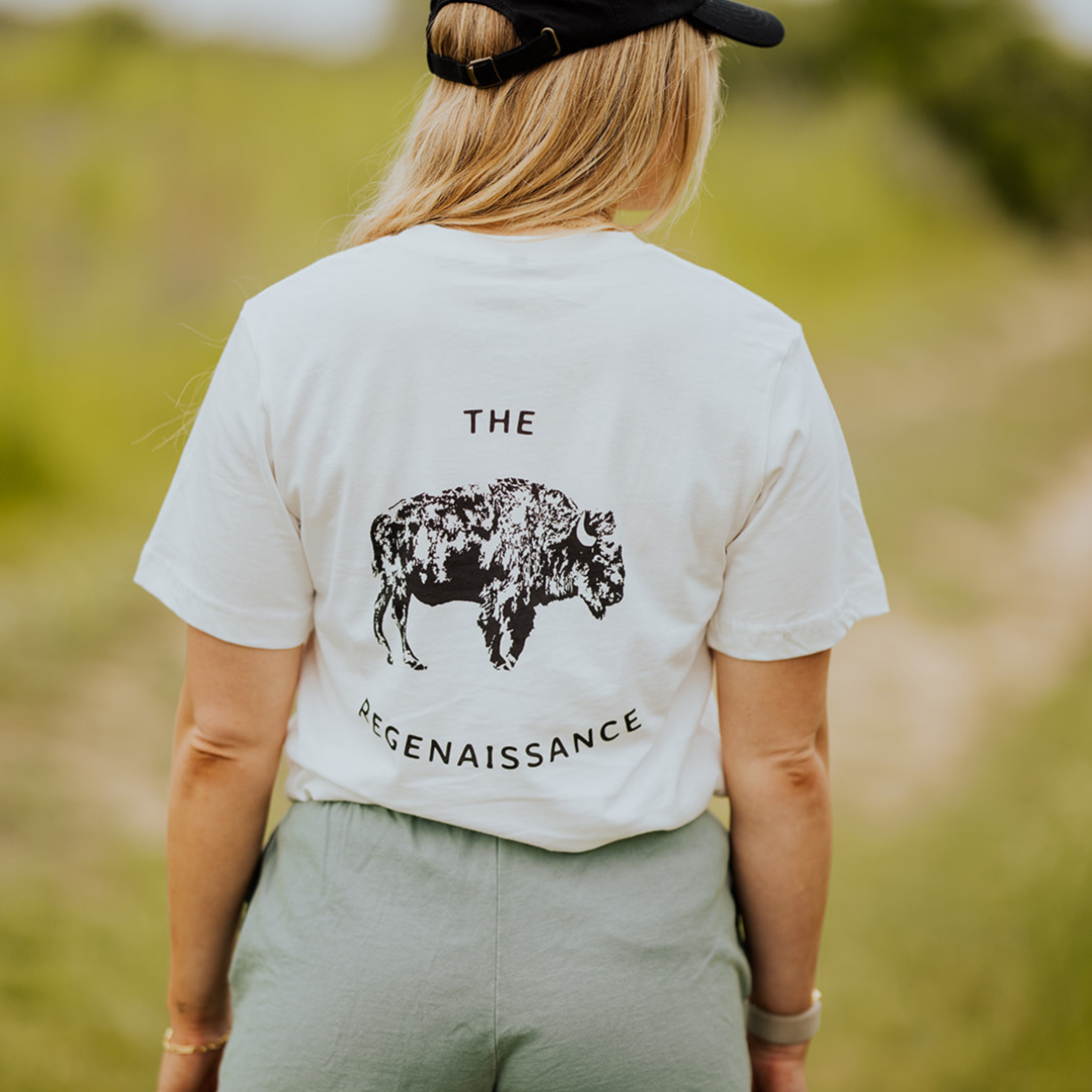 A person stands outdoors on a grassy path, facing away, wearing Los Angeles Apparel Womens Bison Shorts, a black cap, and a white T-shirt with a buffalo graphic and “THE REGENAISSANCE,” referencing regenerative agriculture.