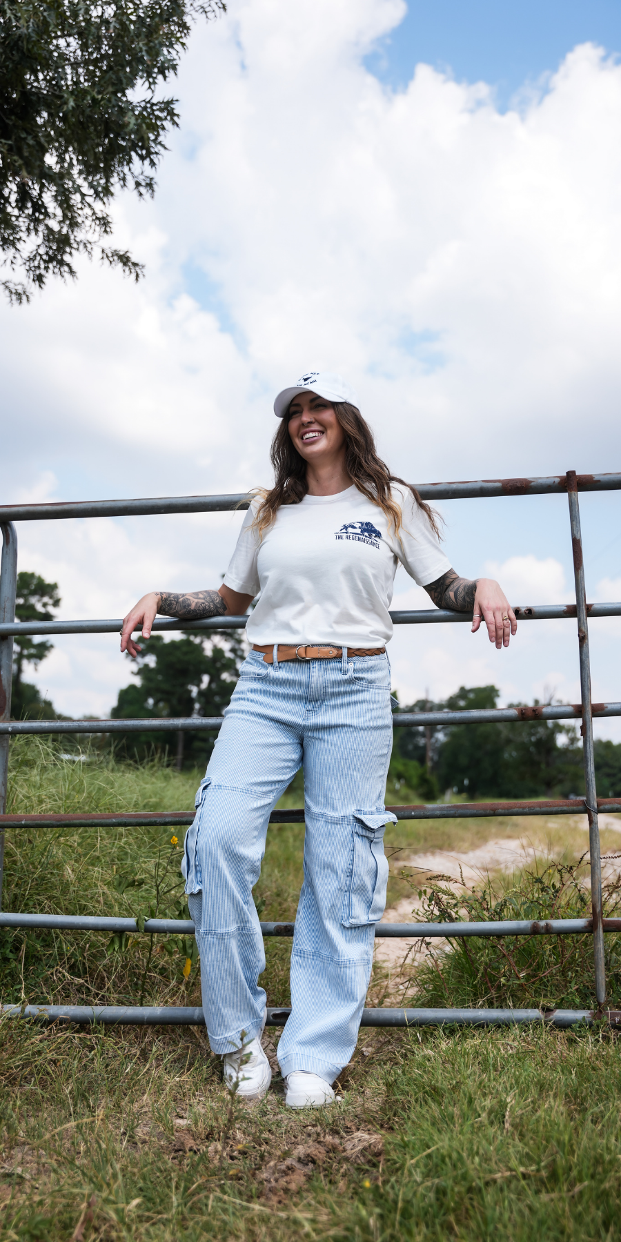A woman stands outdoors leaning against a metal gate, wearing a white graphic t-shirt, light-wash wide-leg cargo jeans, a white baseball cap, and white sneakers.