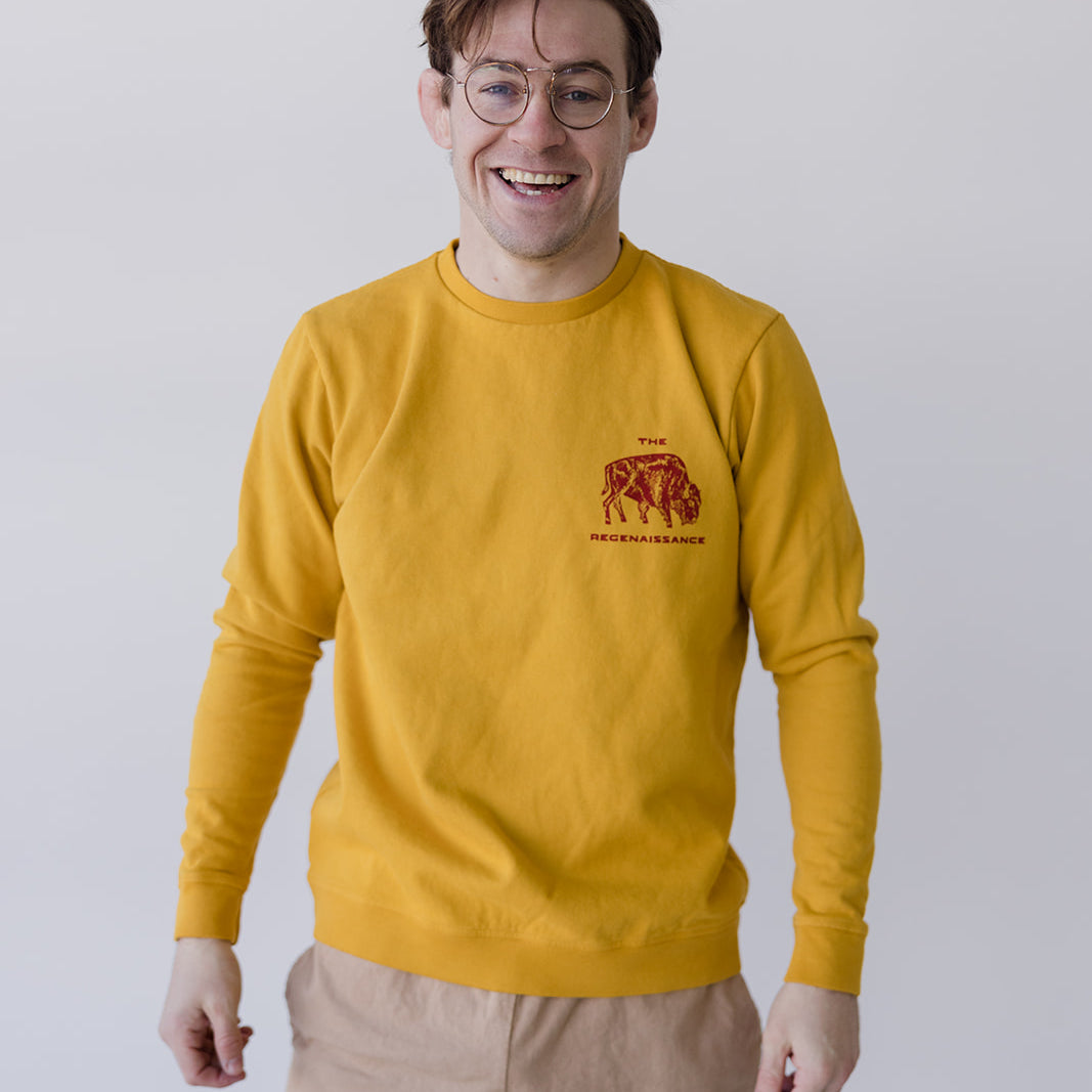 A smiling person in glasses models the Farm Fresh Clothing Bison Crewneck Sweatshirt in mustard and beige shorts with a black design, standing against a plain light background—showcasing sustainable living in style.