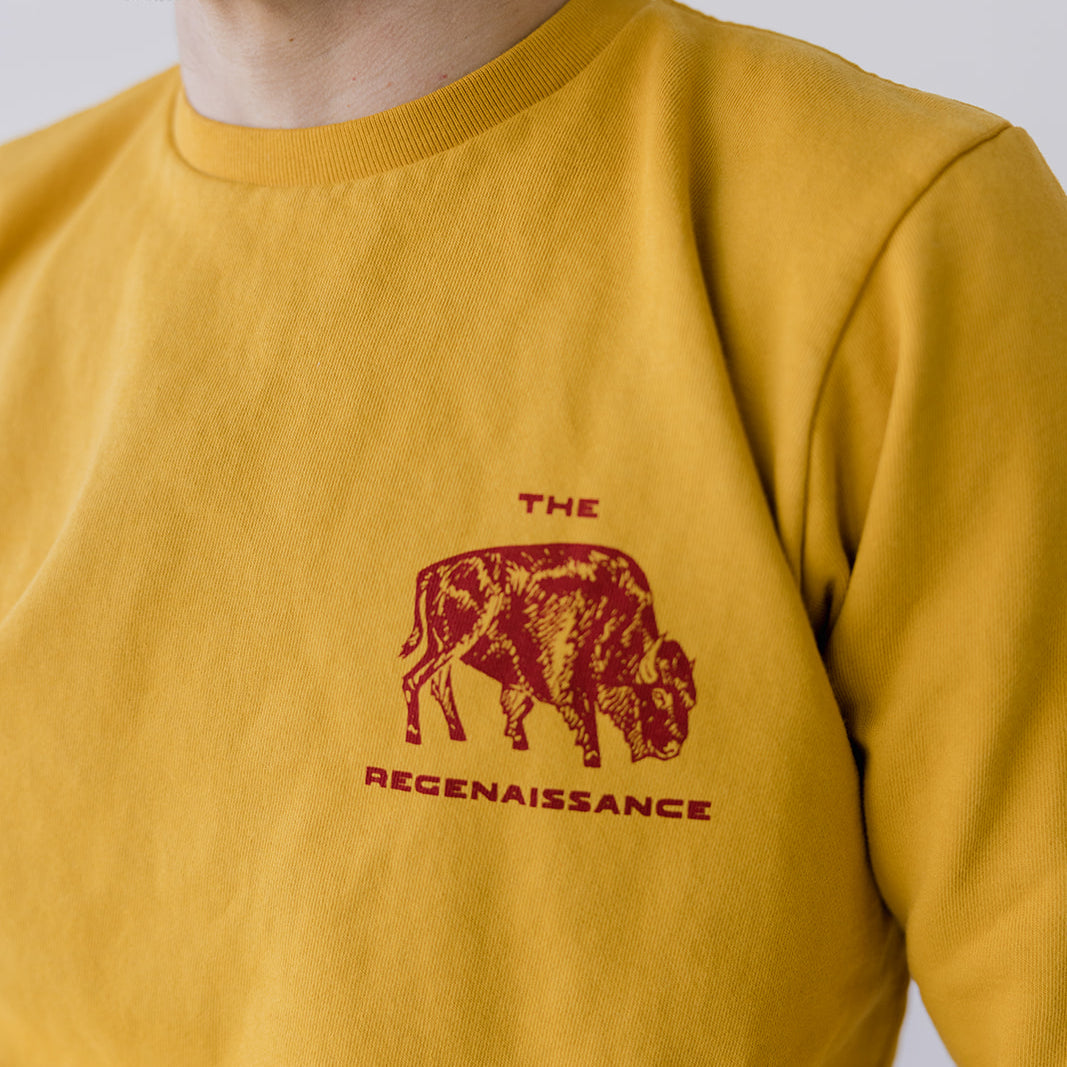 A person wears the Farm Fresh Clothing Bison Crewneck Sweatshirt in mustard yellow organic cotton, featuring a red bison graphic and THE REGENAISSANCE. The face is partially out of frame. Inspired by regenerative agriculture.