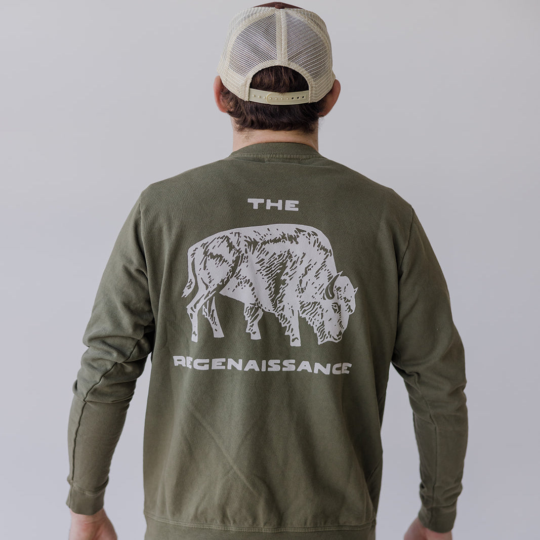 Wearing the Farm Fresh Clothing Bison Crewneck Sweatshirt—green organic cotton with a white bison and THE REGENAISSANCE on the back—and a beige mesh cap, this person stands facing away, embodying sustainable style.