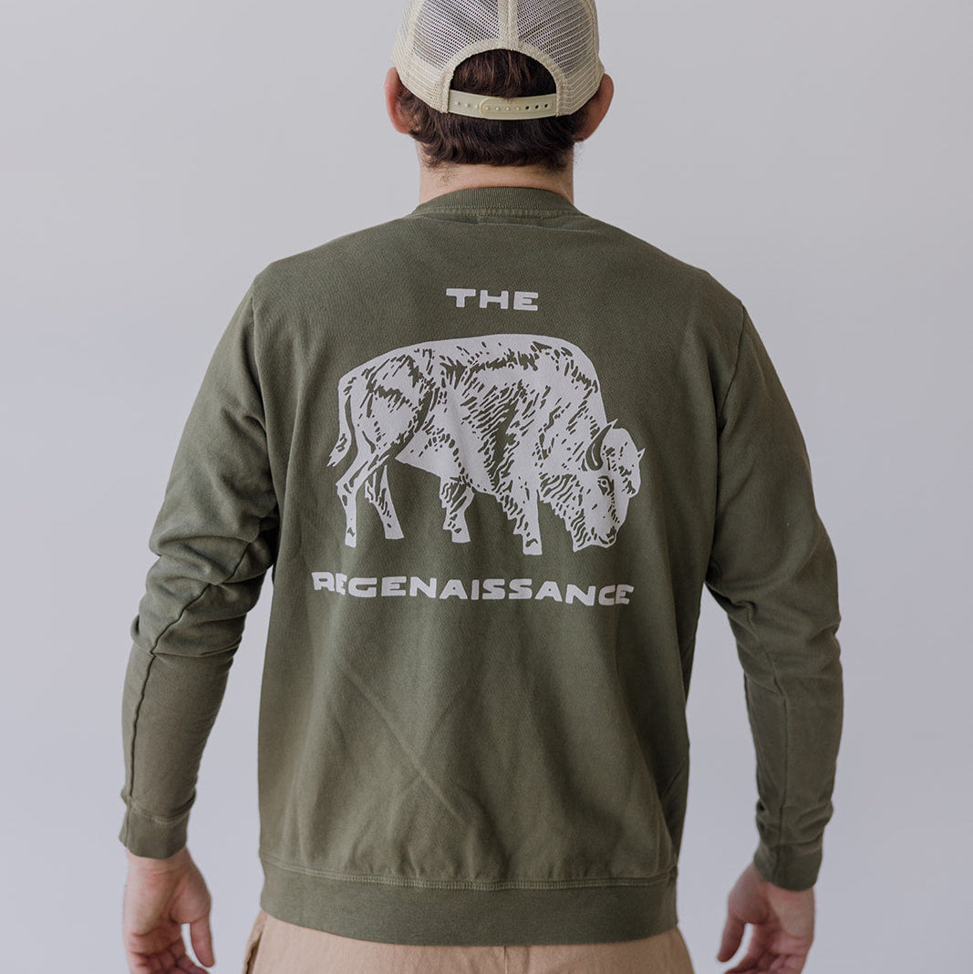 Wearing the Farm Fresh Clothing Bison Crewneck Sweatshirt—green organic cotton with a white bison and THE REGENAISSANCE on the back—and a beige mesh cap, this person stands facing away, embodying sustainable style.